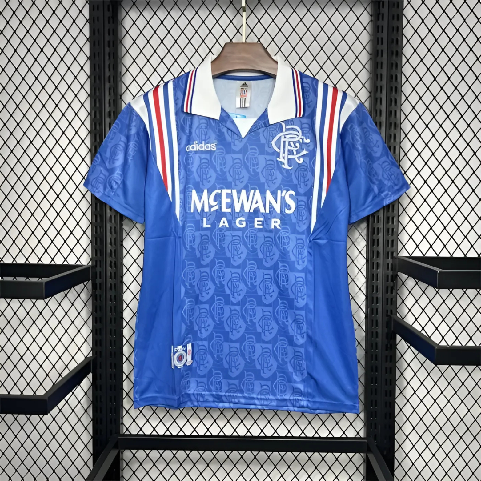 Blue Rangers football jersey with white and red stripes from 1996/97 made by adidas sponsored by McEwan's lager on a hanger in front of a steel fence backdrop