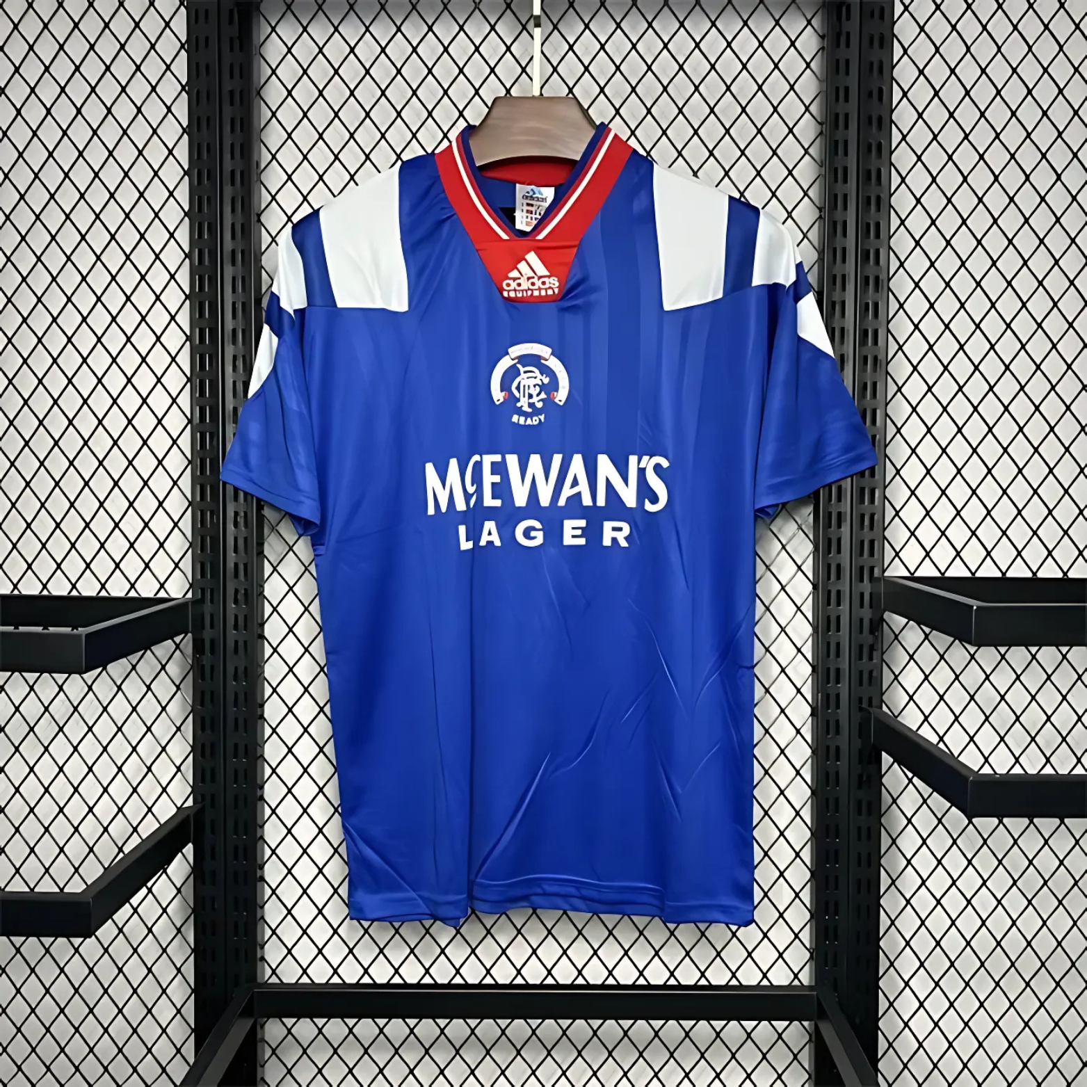 Blue Rangers FC home sports jersey from 1992/94 with 'McEwan's Lager' branding on a hanger against a black metal grid background.