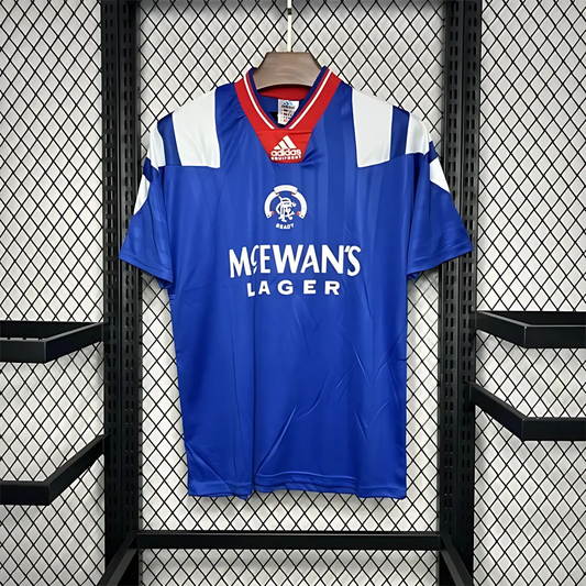 Blue Rangers FC home sports jersey from 1992/94 with 'McEwan's Lager' branding on a hanger against a black metal grid background.